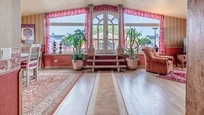 Una habitación con piso de madera, un tapete con diseño rojo y blanco, un banco de madera, una silla rosa y plantas en macetas.