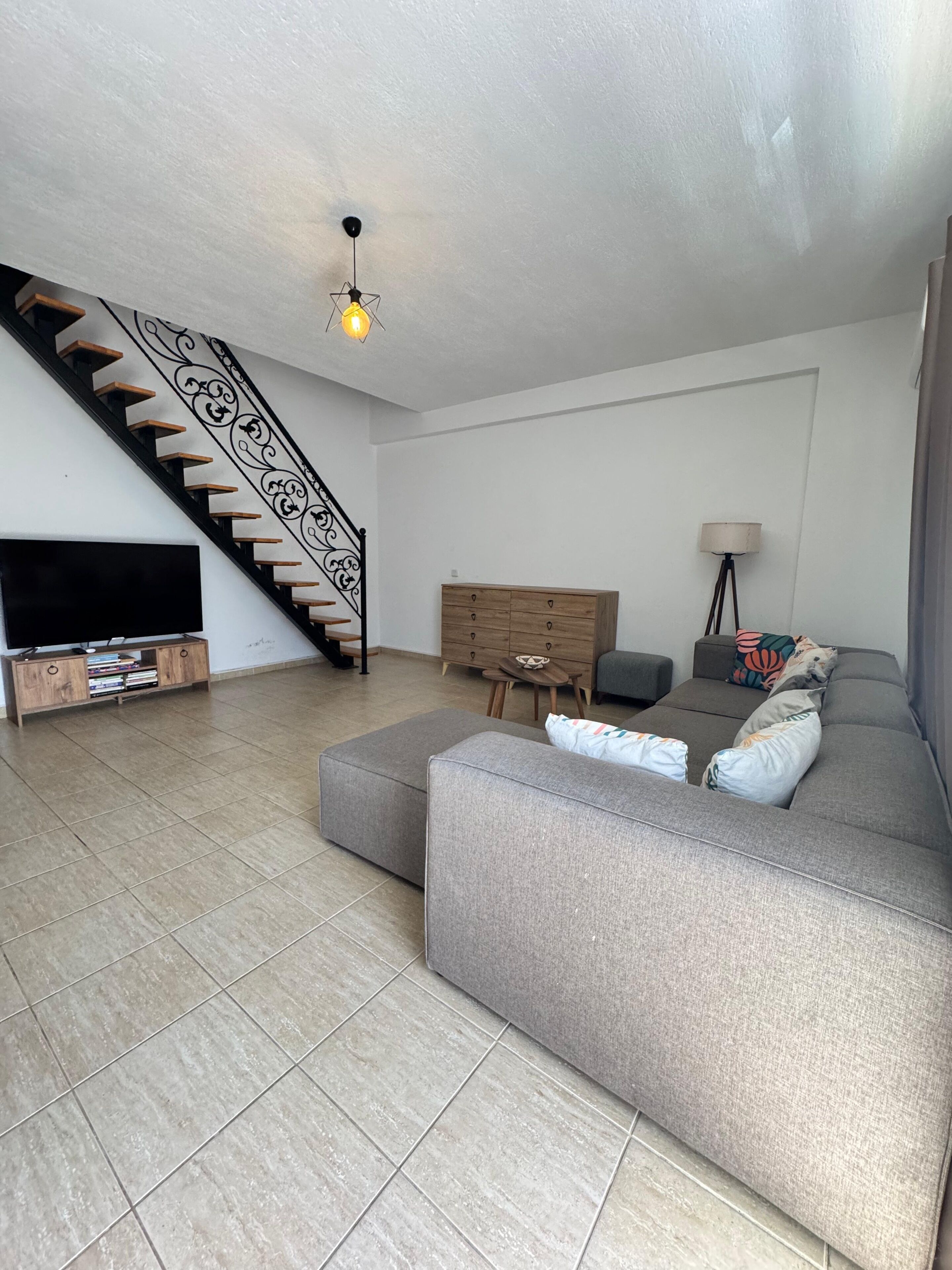 Ein modernes Wohnzimmer mit einem grauen Sofa, einem Holz-TV-Möbel, einer Treppe mit dekorativer Geländer und einer Lampe.