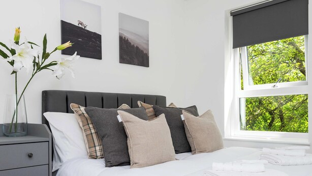 A neatly made bed with white linens, a grey headboard, and multiple pillows in neutral tones. There is a window with a view of green trees, and a vase with white flowers on a bedside table. - Spacious 2BR - Parking Near Edinburgh City Centre (Edinburgh)