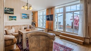 Een woonkamer met een bank, een bijzettafeltje en een televisie. Er zijn grote ramen met gordijnen, een rood gestreepte parasol buiten en kunstwerken aan de muren.