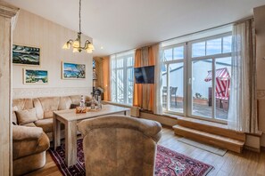 A living room with a sofa, a coffee table, and a TV. There are large windows with curtains, a red-striped umbrella outside, and artwork on the walls. - Wolke 7 Apartment 18 with Sea View and Private Rooftop Terrace (Binz)