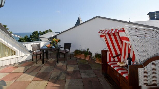 Een terras op het dak met een tafel, stoelen, een rieten bank en potplanten.