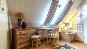 Een kamer met schuine wanden, een houten dressoir, een rieten stoel, een bureau, een radiator en een raam met gordijnen.