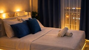 Siististi pedattu sänky, jossa on valkoinen patja ja siniset tyynyt, yöpöytä lampun kanssa ja ikkuna, jossa on verhot.