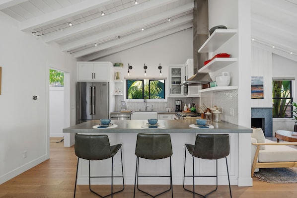 A modern kitchen with a central island, bar stools, and a view of greenery outside.