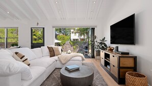 A modern living room with a white sofa, a round coffee table, a flat-screen TV, and a view of a garden through large windows. - Humphrey House | Walk to Beach, Coast Village Rd, 3 En Suite Bedrooms (Montecito)
