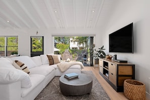 A modern living room with a white sofa, a round coffee table, a flat-screen TV, and a view of a garden through large windows.
