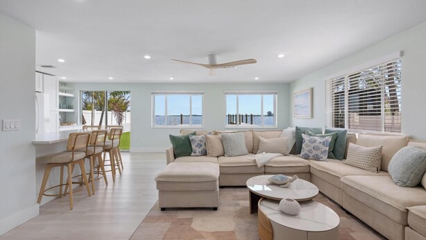 A modern living room with a sectional sofa, a dining area with bar stools, and large windows with a view of the sea. - Manatee Cove -3-bedroom bayfront home with dock (Bradenton Beach)
