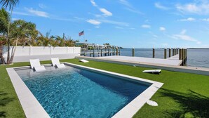 A rectangular swimming pool with clear blue water, surrounded by artificial grass and white lounge chairs. - Manatee Cove -3-bedroom bayfront home with dock (Bradenton Beach)