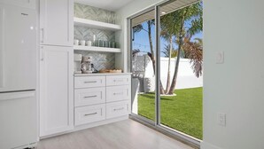 A modern kitchen with white cabinetry, a built-in coffee maker, and a view of a well-maintained lawn and palm trees through a sliding glass door. - Manatee Cove -3-bedroom bayfront home with dock (Bradenton Beach)