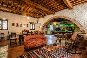 Un interno rustico con soffitto in legno, un divano decorato, una zona pranzo e una cucina visibile attraverso un arco.