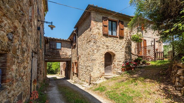 A narrow stone pathway leading to a rustic stone building with wooden shutters and a small porch with potted plants.