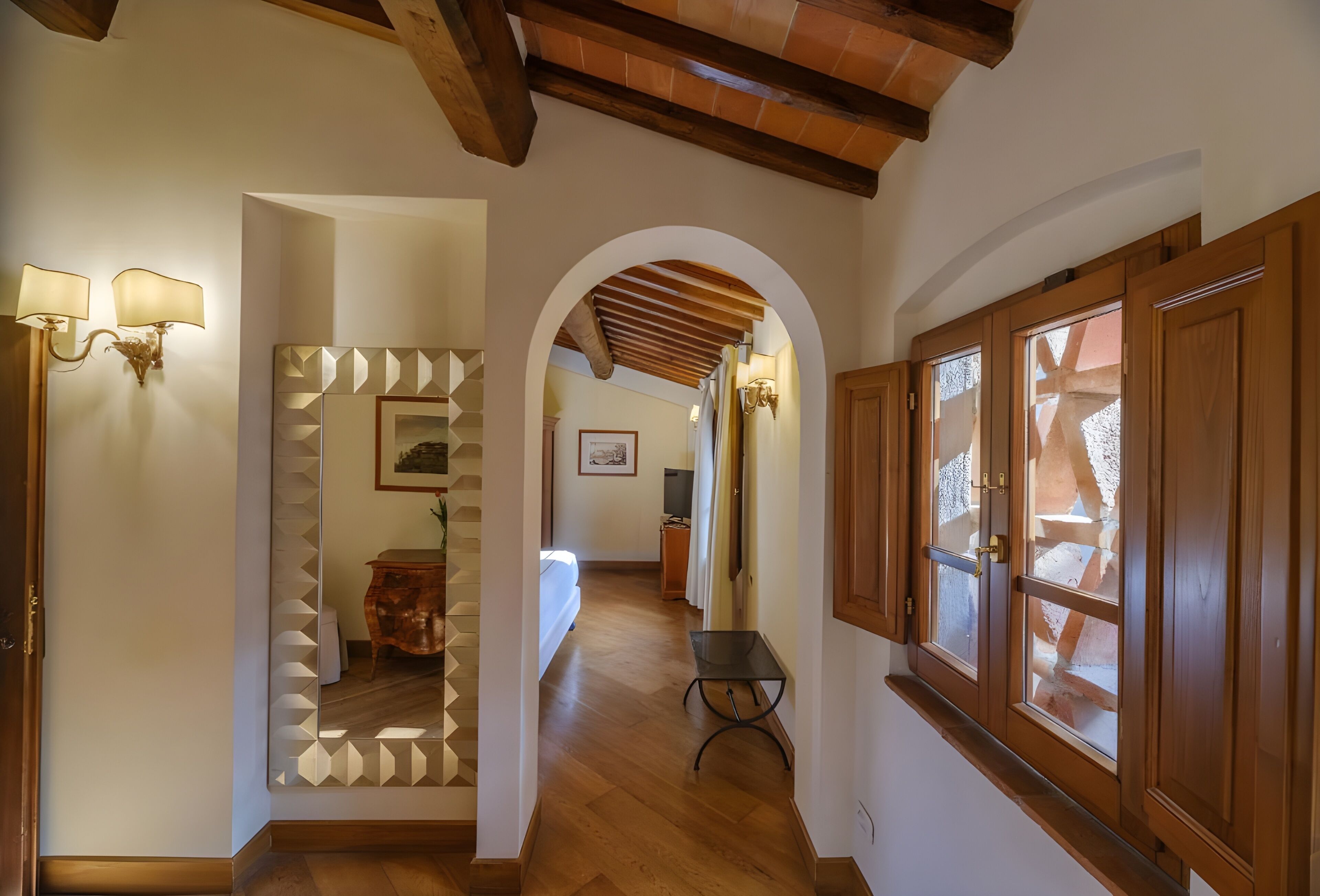 Un pasillo con suelo de madera, un arco de entrada, un espejo con diseños geométricos y una cama al fondo.