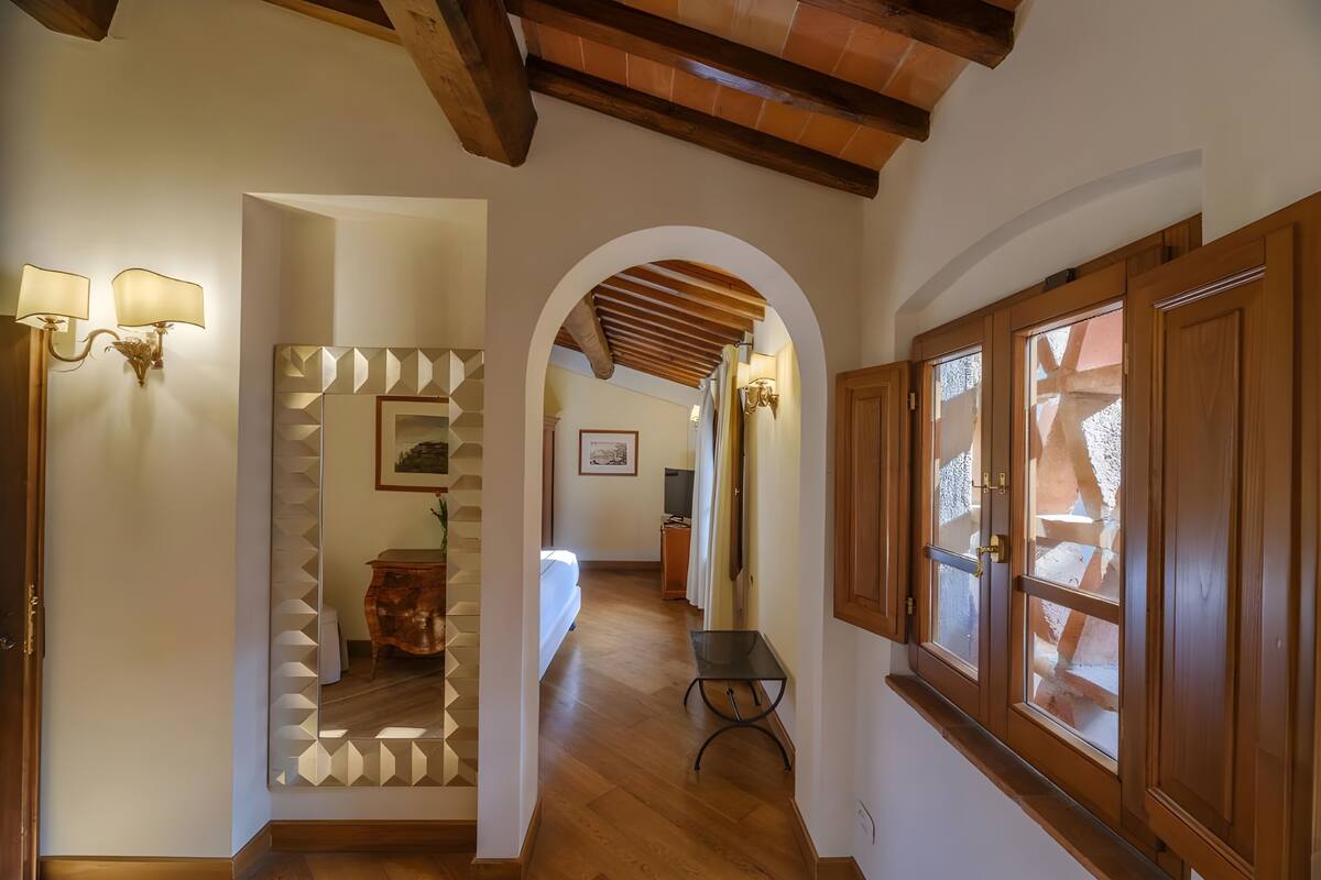 Un pasillo con suelo de madera, un arco de entrada, un espejo con diseños geométricos y una cama al fondo.