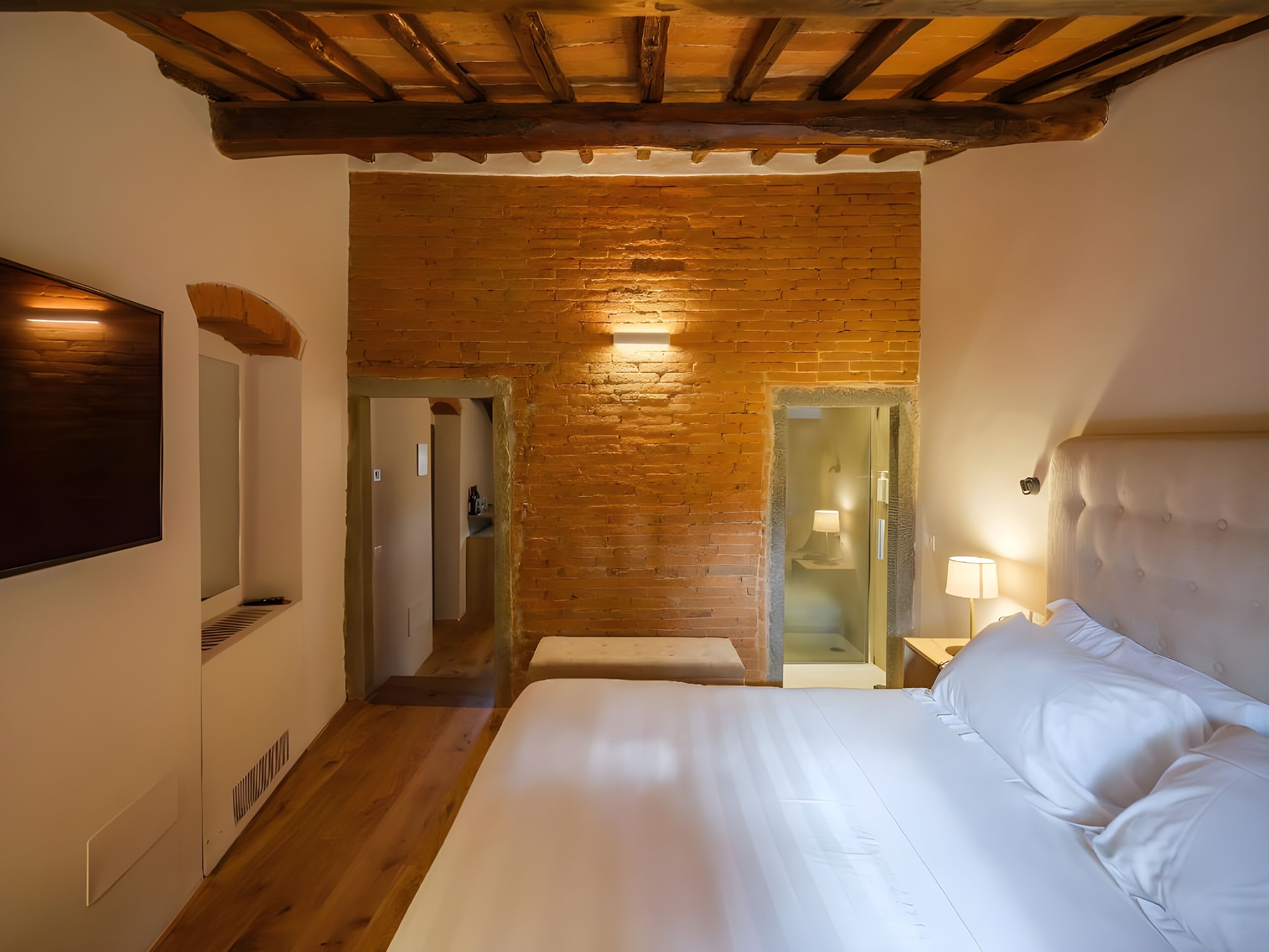 Un dormitorio con techo de madera, una cama grande, un televisor fijado a la pared y un baño visible a través de una puerta de cristal.