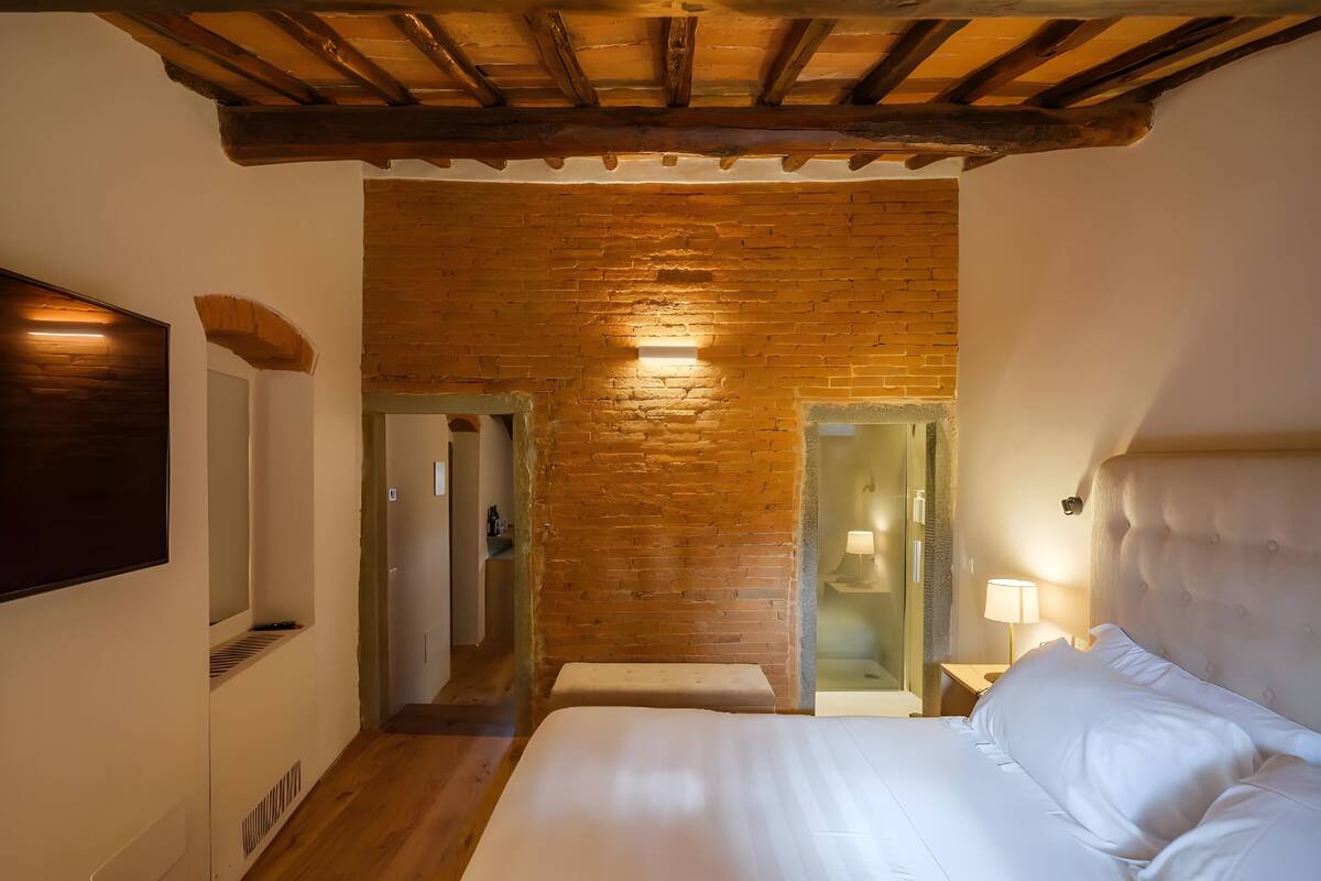 Un dormitorio con techo de madera, una cama grande, un televisor fijado a la pared y un baño visible a través de una puerta de cristal.