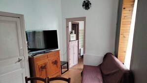 Ein Zimmer mit Fernseher auf einem Holzkasten, einem Esstisch mit Stühlen, einem violetten Sofa und einer Tür zu einem anderen Raum.