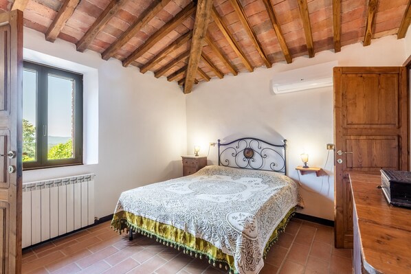 Een slaapkamer met een houten plafond, een bed met een bedsprei met dessin, een nachtkastje met een lamp, een raam met uitzicht en een radiator.