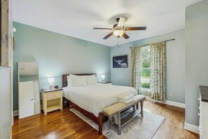 Un dormitorio con cama, mesitas de noche, ventilador de techo, ventana con cortinas y vistas al exterior.