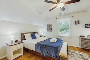 Un dormitorio con cama, mesitas de noche, ventilador de techo, ventana con persianas y un pequeño escritorio con cajones.