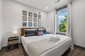 A neatly arranged bedroom with a bed, bedside table, and a window with a view of trees. - Music City Condo・Mins to Broadway W Rooftop Lounge (Nashville)