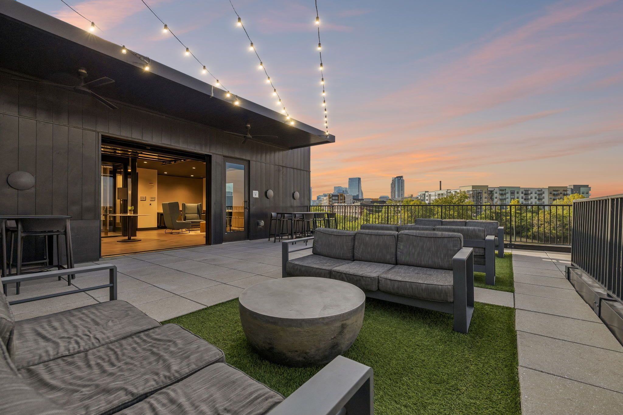 An outdoor seating area with a modern design, featuring sofas and a round table, set against a cityscape view at sunset.