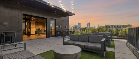 An outdoor seating area with a modern design, featuring sofas and a round table, set against a cityscape view at sunset.