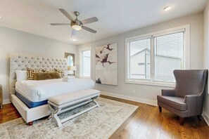 A bedroom with a tufted headboard, a bed with white bedding, a bench, a chair, a ceiling fan, and artwork on the walls. - Brand-New Spacious Modern House with Rooftop Deck & City Views (Nashville)