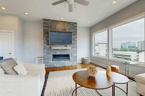 A modern living room with a stone fireplace, a flat-screen TV, a large window with a city view, and a round coffee table. - Brand-New Spacious Modern House with Rooftop Deck & City Views (Nashville)