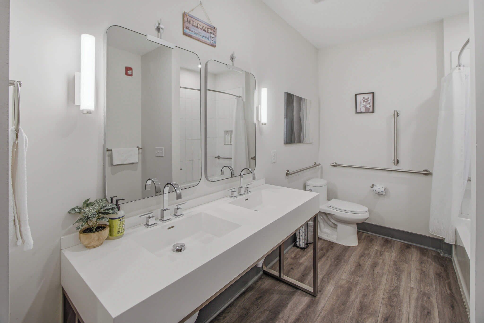 A modern bathroom with a double sink, a toilet, a shower, and a handrail.