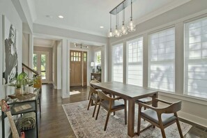 A spacious dining area with a long wooden table and chairs, large windows with blinds, and a staircase leading to another room.