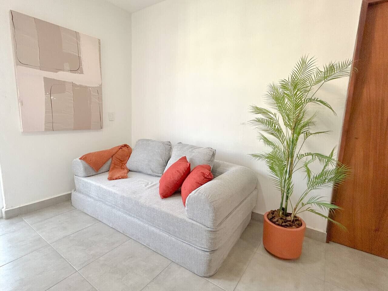 Ein graues Sofa mit roten und grauen Kissen, ein Blumentopf und eine weiße Wand mit einem großen abstrakten Gemälde.