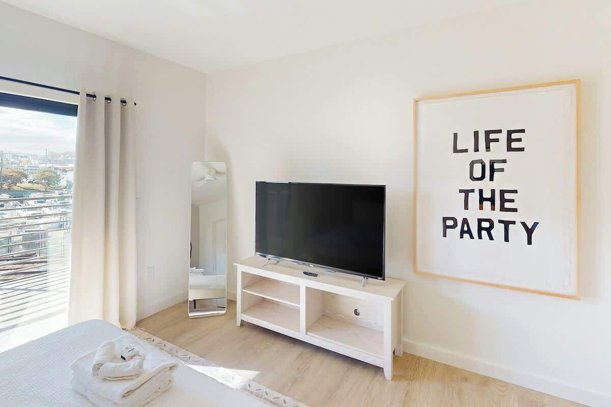 Une chambre d’hôtel avec un lit, un téléviseur posé sur un meuble blanc, un miroir et un tableau encadré accroché au mur.