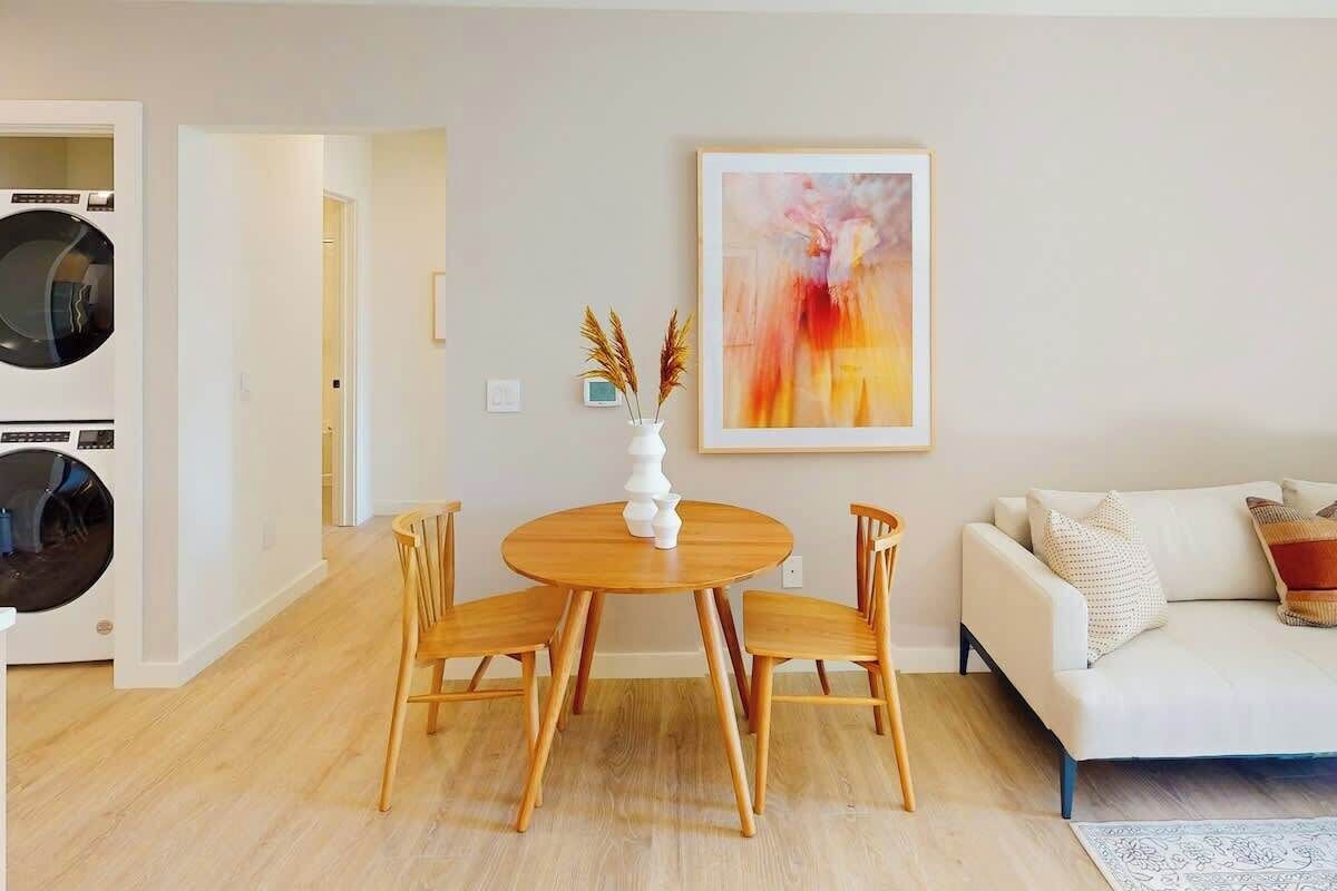A living room with a round wooden table, four wooden chairs, a white sofa, and a painting on the wall.