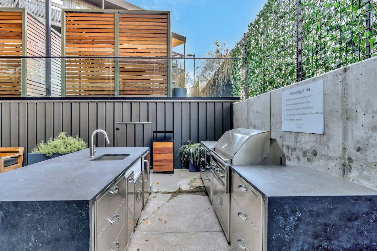 Cocina exterior con pileta, parrilla y armarios, frente a un edificio moderno y un seto verde.