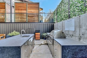 An outdoor kitchen with a sink, grill, and cabinets, set against a modern building and a green hedge. - Centrally located condo with pool & gym access, balcony, WD & Modern Interior (Nashville)