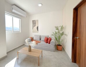 A living room with a grey sofa, a wooden coffee table, a potted plant, and a large window. - Estancia Cómoda en Centro Vallarta - 3 (Puerto Vallarta)