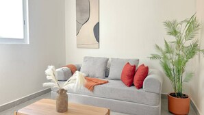Un salon avec un canapé gris, des coussins rouges, une table basse en bois, un vase avec des plumes blanches, une plante en pot et une œuvre d’art murale.