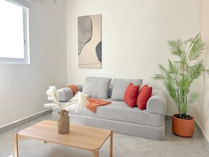 A living room with a grey sofa, red cushions, a wooden coffee table, a vase with white feathers, a potted plant, and a wall art piece. - Estancia Cómoda en Centro Vallarta - 3 (Puerto Vallarta)