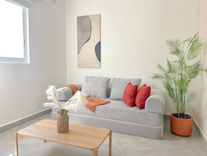 Ein Wohnzimmer mit grauem Sofa, roten Kissen, einem Holztisch, einer Vase mit weißen Federn, einer Topfpflanze und einem Wandbild.