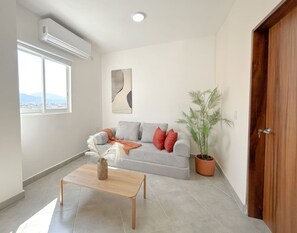 A living room with a grey sofa, a wooden coffee table, a potted plant, and a large window. - Estancia Cómoda en Centro Vallarta - 5 (Puerto Vallarta)