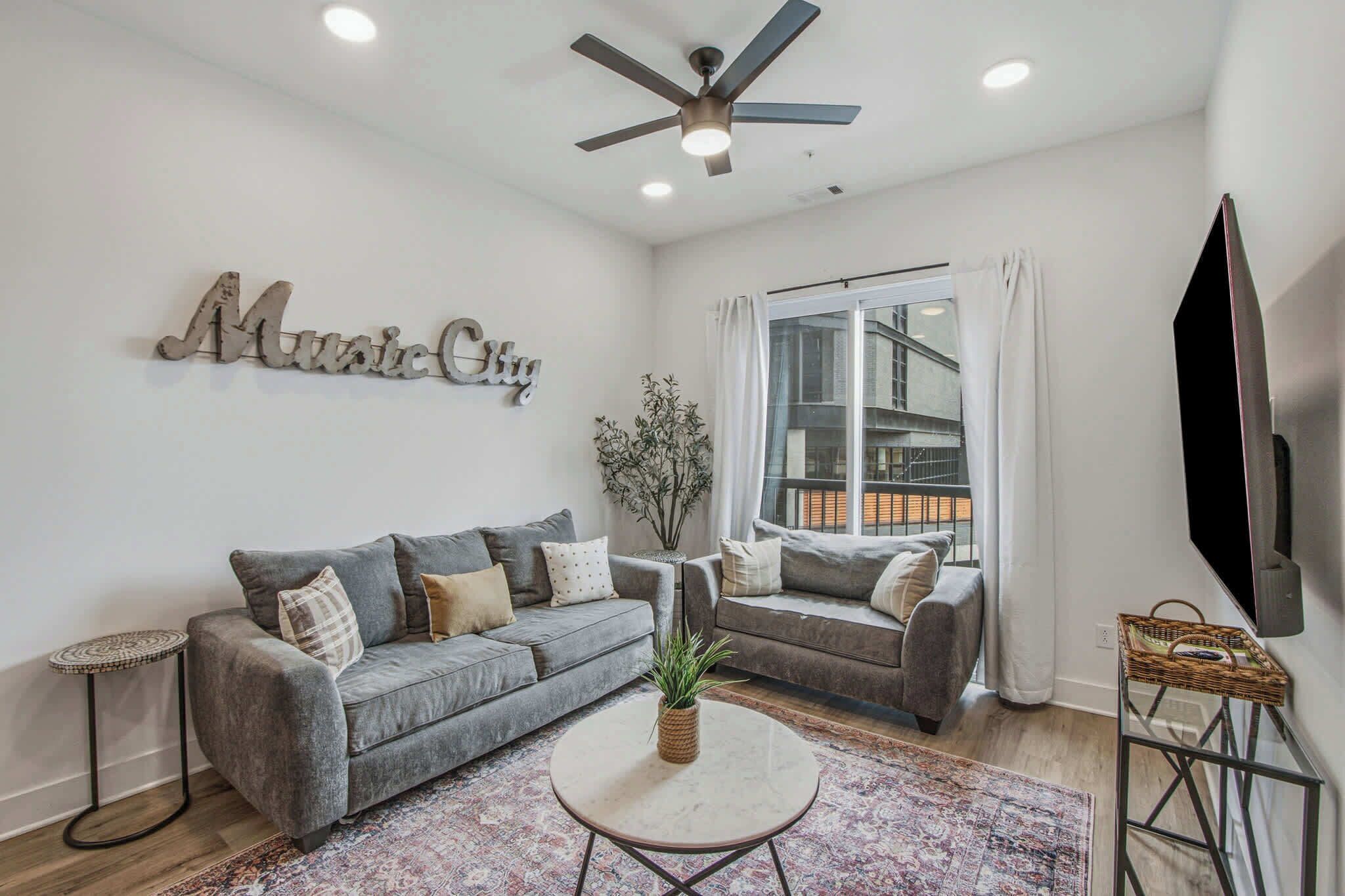 A living room with a grey sofa, a coffee table, a plant, and a wall-mounted TV.