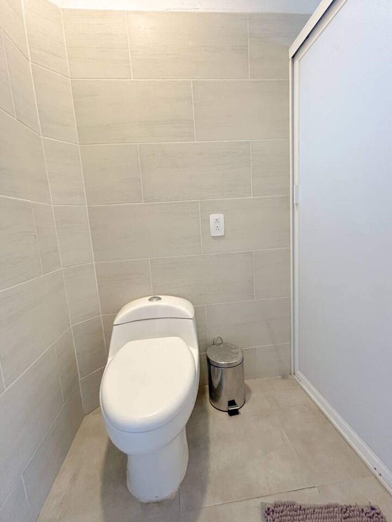 A small, clean bathroom with a toilet, a trash can, and a power outlet on the wall.