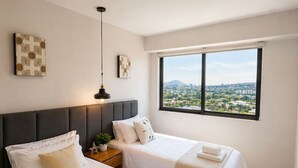 Ein Hotelzimmer mit zwei Betten, einem großen Fenster mit Blick auf die Stadt, einer Pendelleuchte und Kunstwerken an den Wänden.
