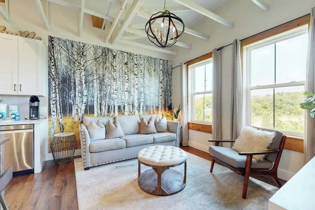 Un salon moderne avec un canapé, un pouf et un fauteuil, une grande fenêtre avec des rideaux et une tapisserie murale sur le thème de la forêt.