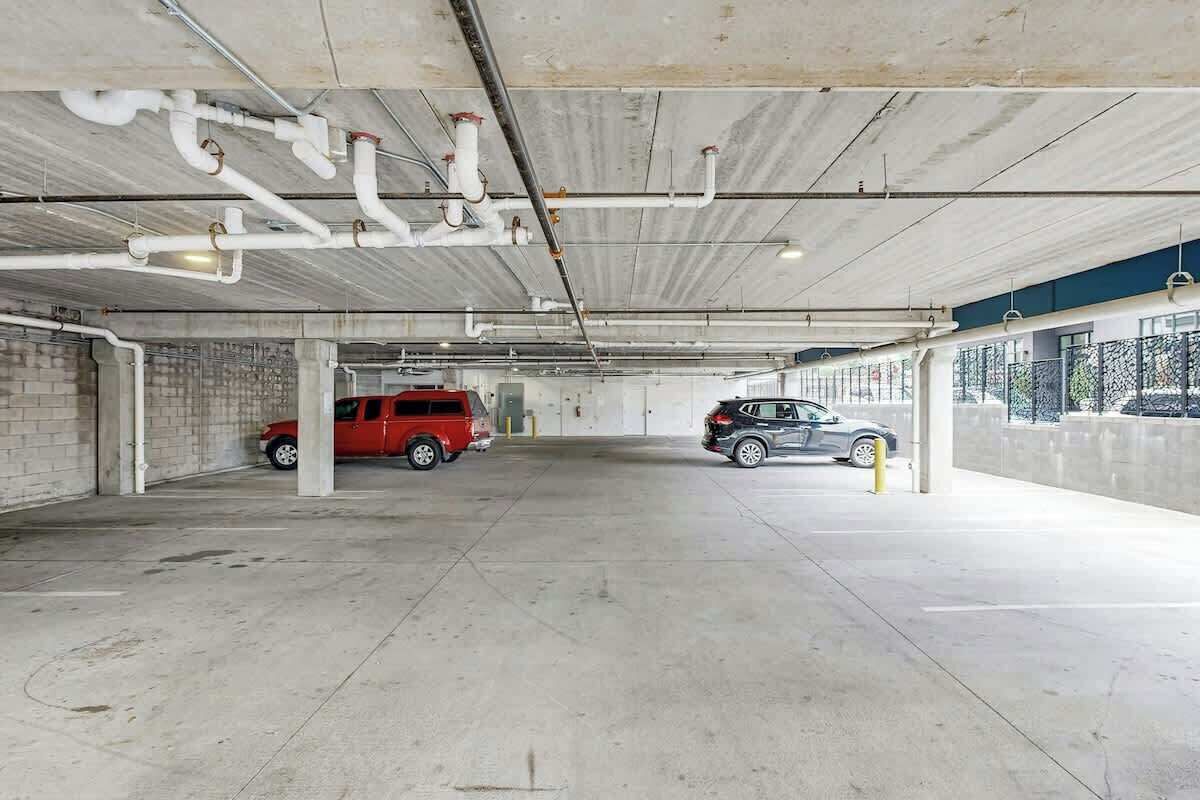 En parkeringskælder med betongulv og -vægge, synlige rør og kanaler i loftet, og to parkerede biler.