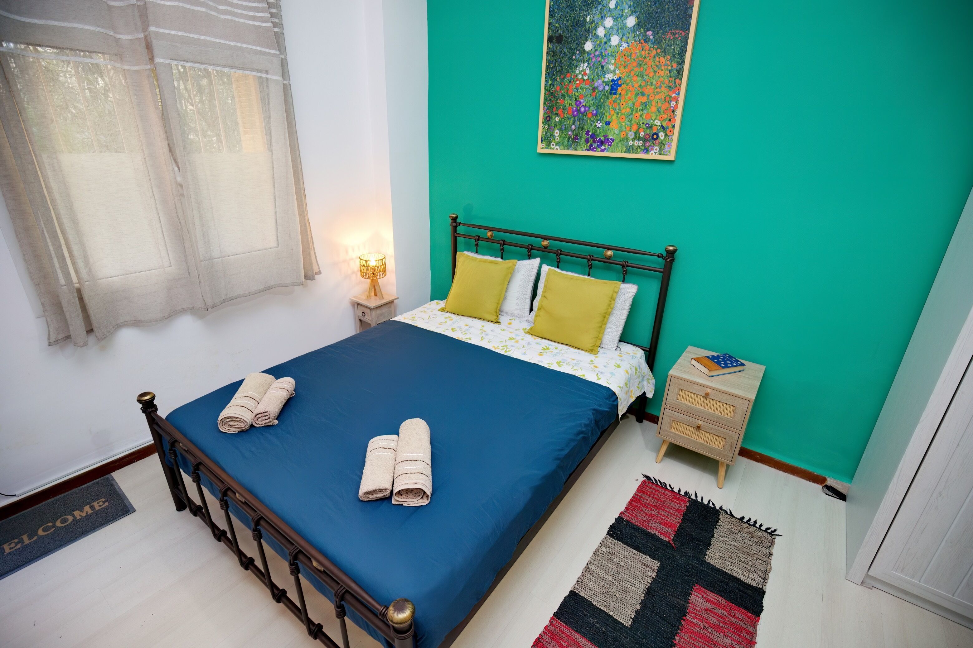 Een keurig opgemaakt bed met blauw beddengoed en twee opgerolde handdoeken, een nachtkastje met een lamp, een klein houten nachtkastje, een kleurrijk abstract schilderij aan de muur, en een deurmat bij de deur.