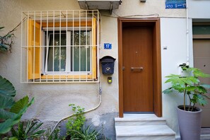 A building with a wooden door, a window with yellow shutters, and a mailbox. - Under Lycabettus Hideaway by ΜΡS (Athina)