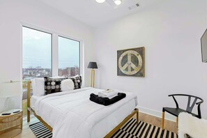 Ein modernes Schlafzimmer mit großem Fenster, einem Bett mit weißer Bettwäsche, einem schwarz-weiß gestreiften Teppich und einem schwarzen Stuhl.