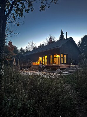 A wooden cabin with a porch, a chimney, and a view of the surrounding forest.